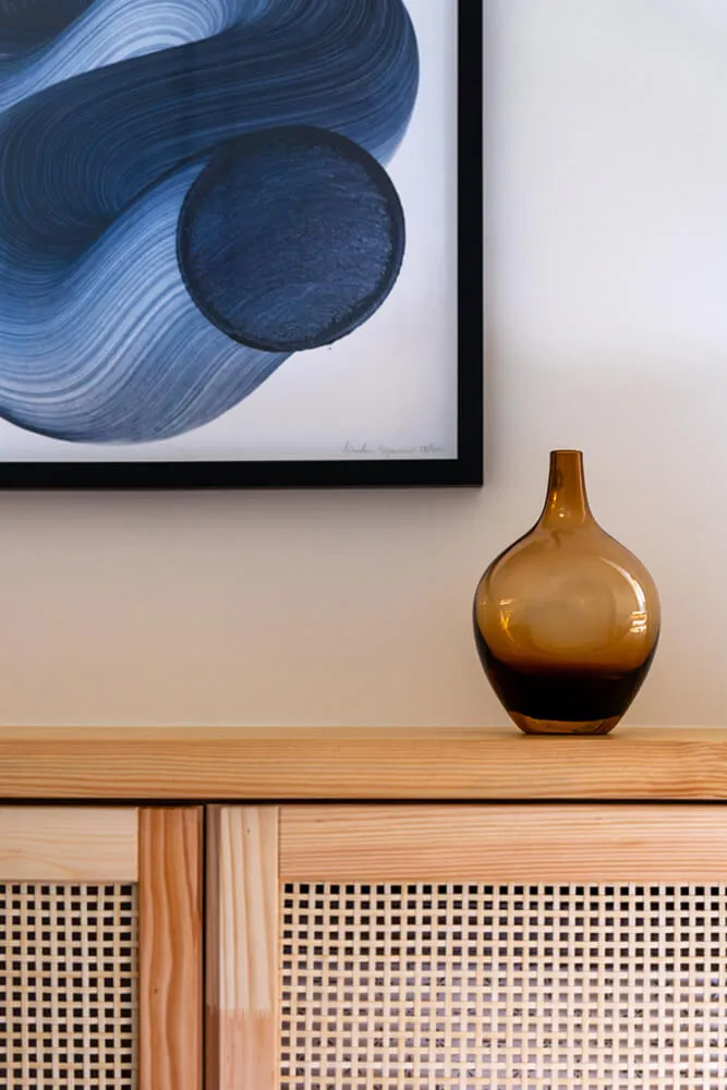Brązowa przezroczysta wazonowa na drewnianym kredensie pod abstrakcyjnym obrazem z niebieskimi i szarymi okręgami.