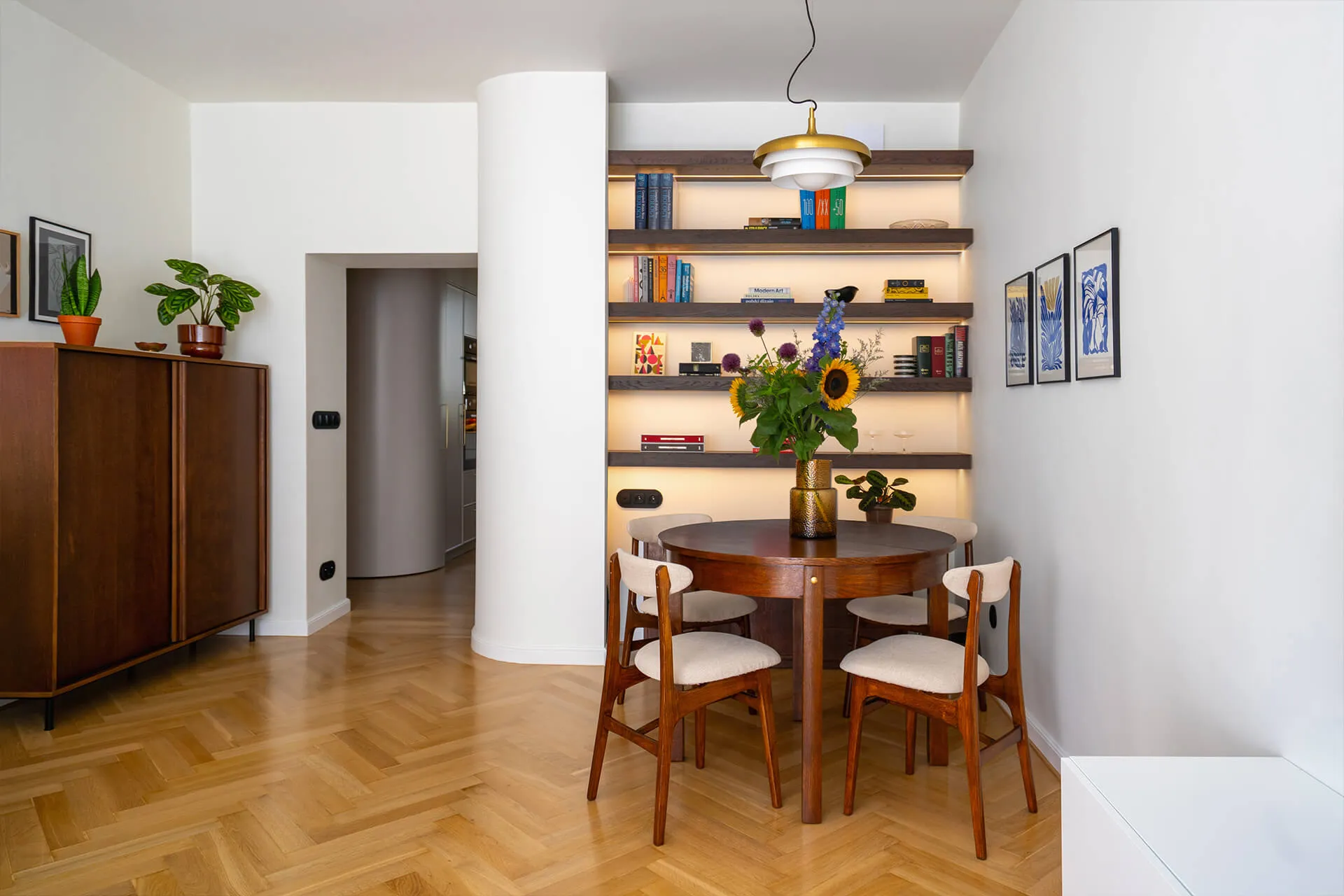 Jadalnia z okrągłym drewnianym stołem i czterema krzesłami, na stole wazon z kwiatami, podświetlane półki z książkami na ścianie i komoda z roślinami w doniczkach po lewej.