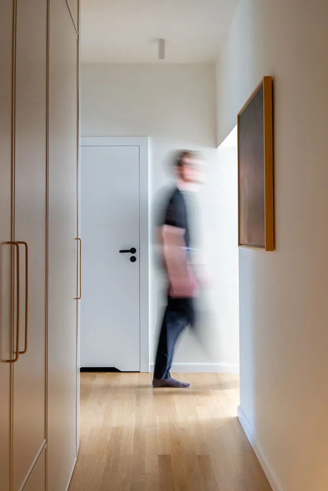 Rozmyta sylwetka osoby chodzącej po jasnym korytarzu z drewnianą podłogą, białymi ścianami i szafkami po lewej stronie.