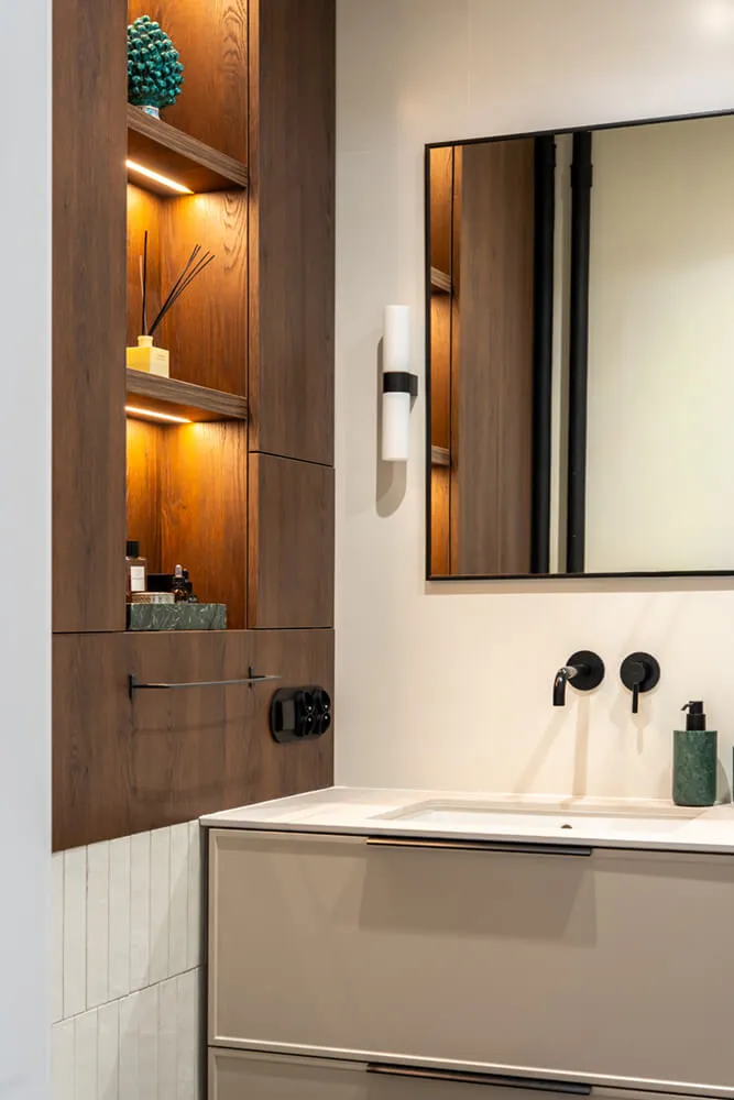 Nowoczesna łazienka z jasną umywalką, czarnym lustrem, oświetleniem i drewnianymi półkami z dekoracjami.