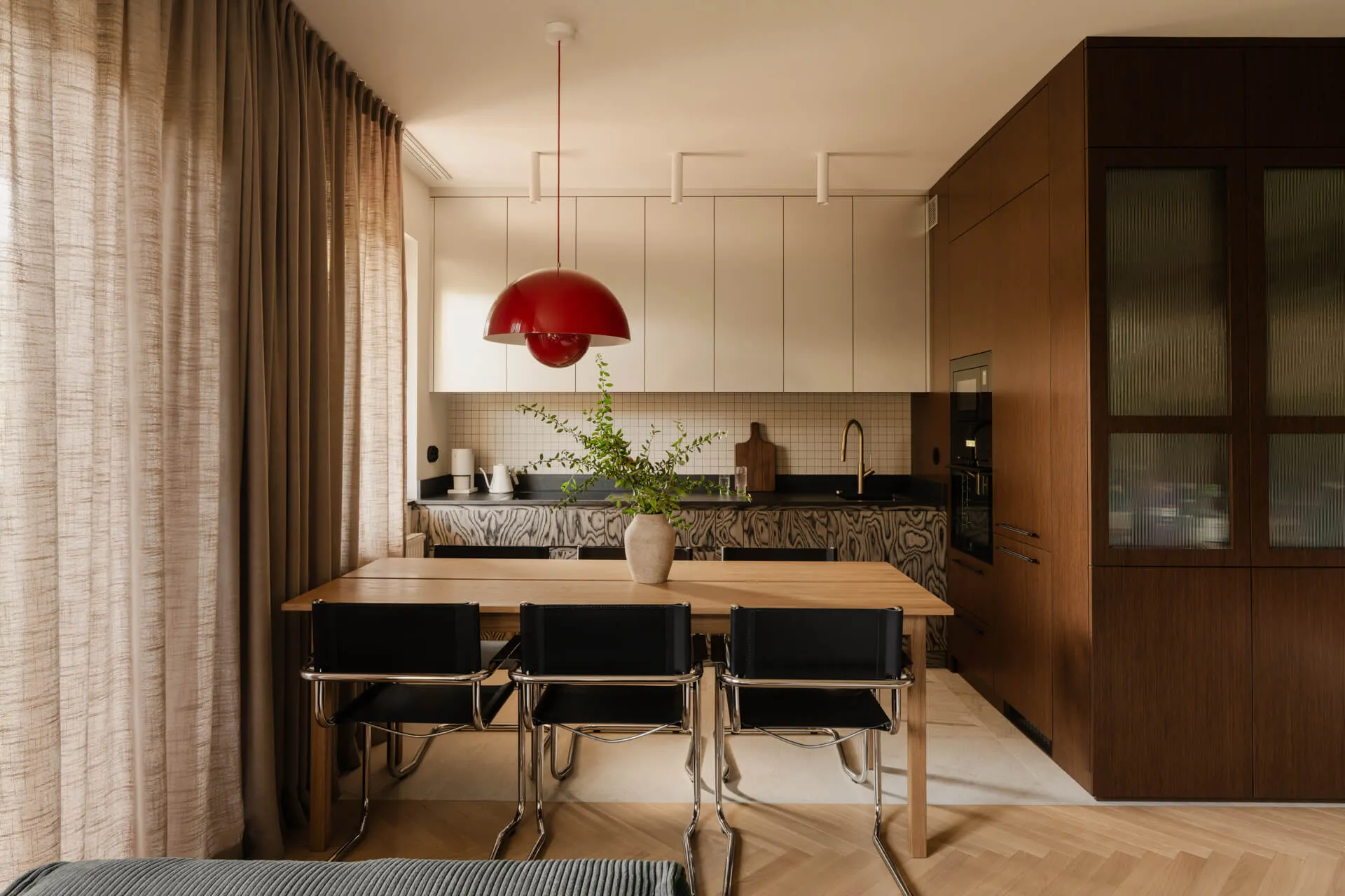 Nowoczesna kuchnia z drewnianym stołem, trzema czarnymi krzesłami, czerwoną lampą wiszącą i doniczką z zielonymi roślinami.
