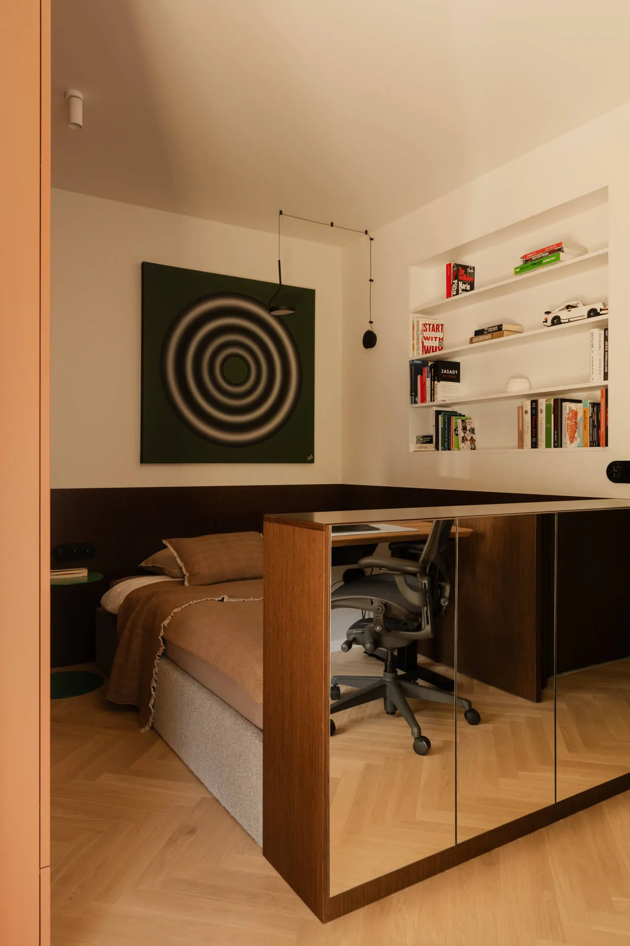 Pokój z łóżkiem w brązowej pościeli, biurkiem z czarnym krzesłem obrotowym, półkami z książkami i obrazem z koncentrycznymi kołami na ścianie.