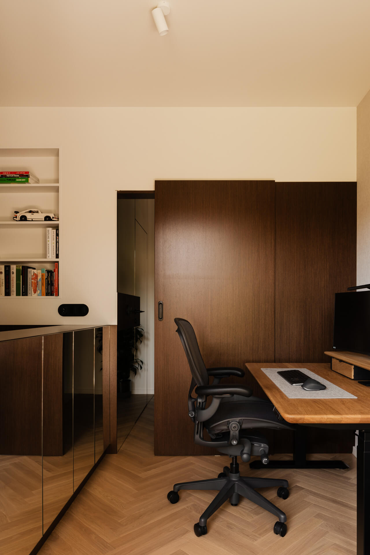 Nowoczesne biuro domowe z czarnym ergonomicznym krzesłem obrotowym, drewnianym biurkiem i ciemnobrązowymi drzwiami przesuwnymi.