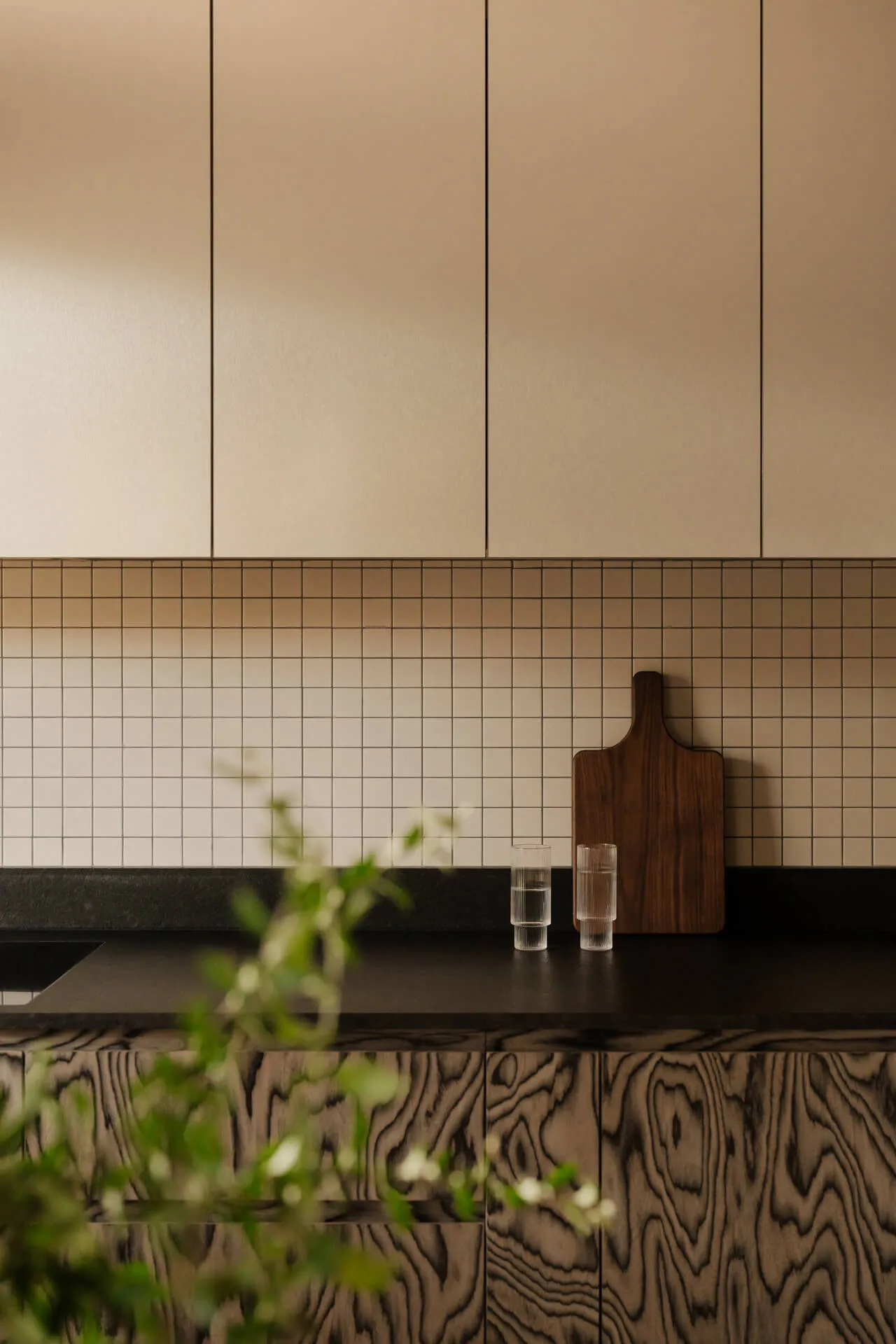 Nowoczesna kuchnia z czarnym blatem, drewnianymi szafkami, jasnymi górnymi szafkami, płytkami w kratkę na ścianie, deską do krojenia i dwoma szklankami z wodą.