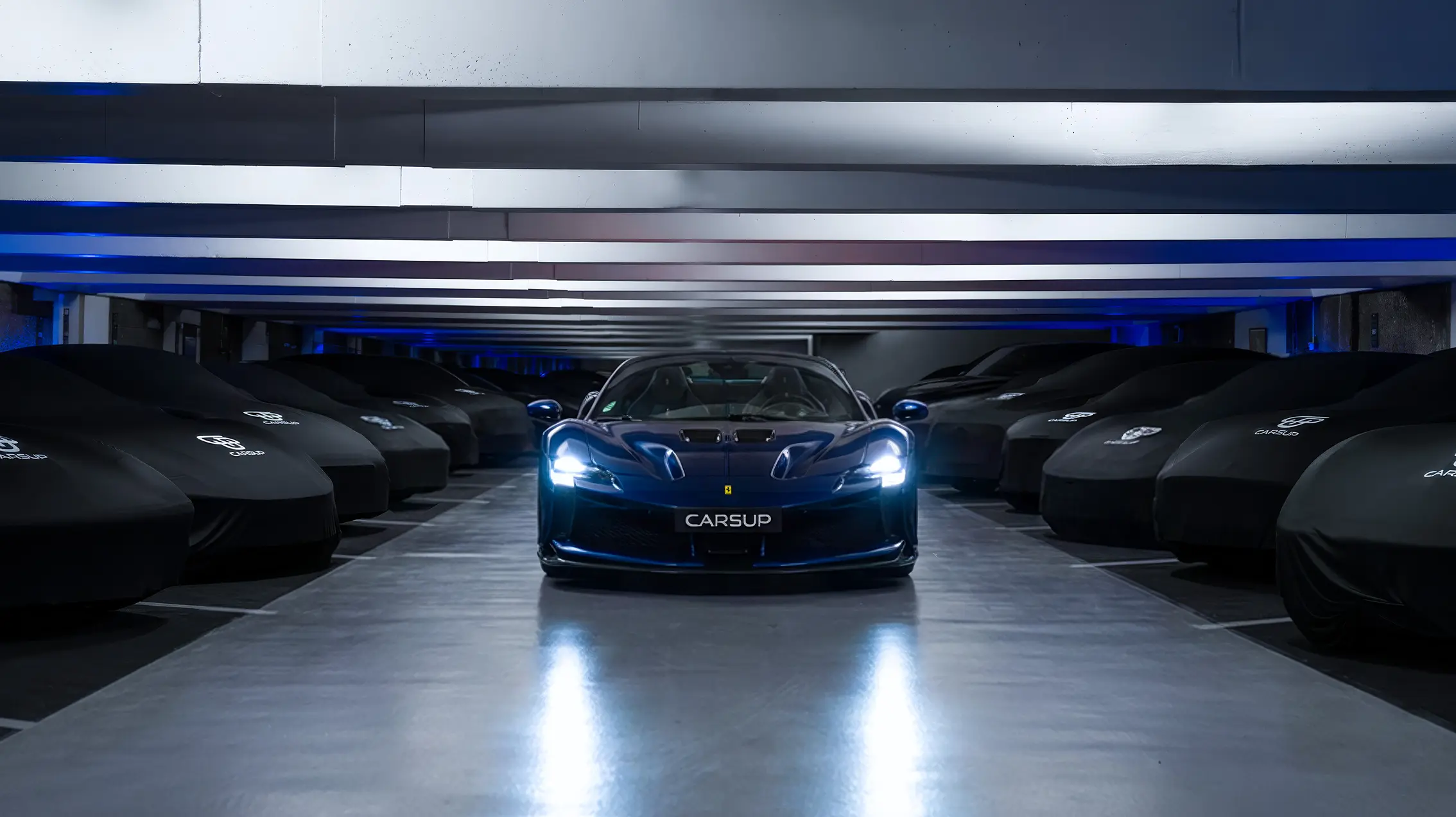 Luxury blue Ferrari centered in a high-end underground garage, headlights on, surrounded by covered sports cars on both sides under sleek modern lighting, with a “CARSUP” plate on the front.