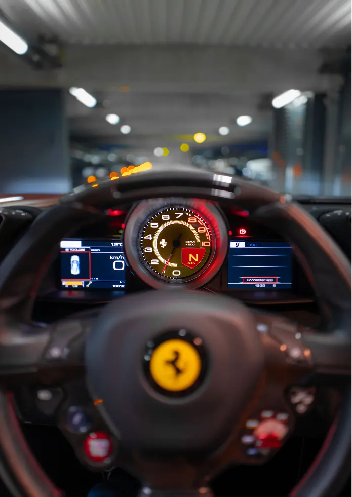 Driver’s perspective inside a Ferrari, focused on the illuminated central tachometer and digital dashboard, with the steering wheel and prancing horse logo in the foreground inside a modern parking garage.