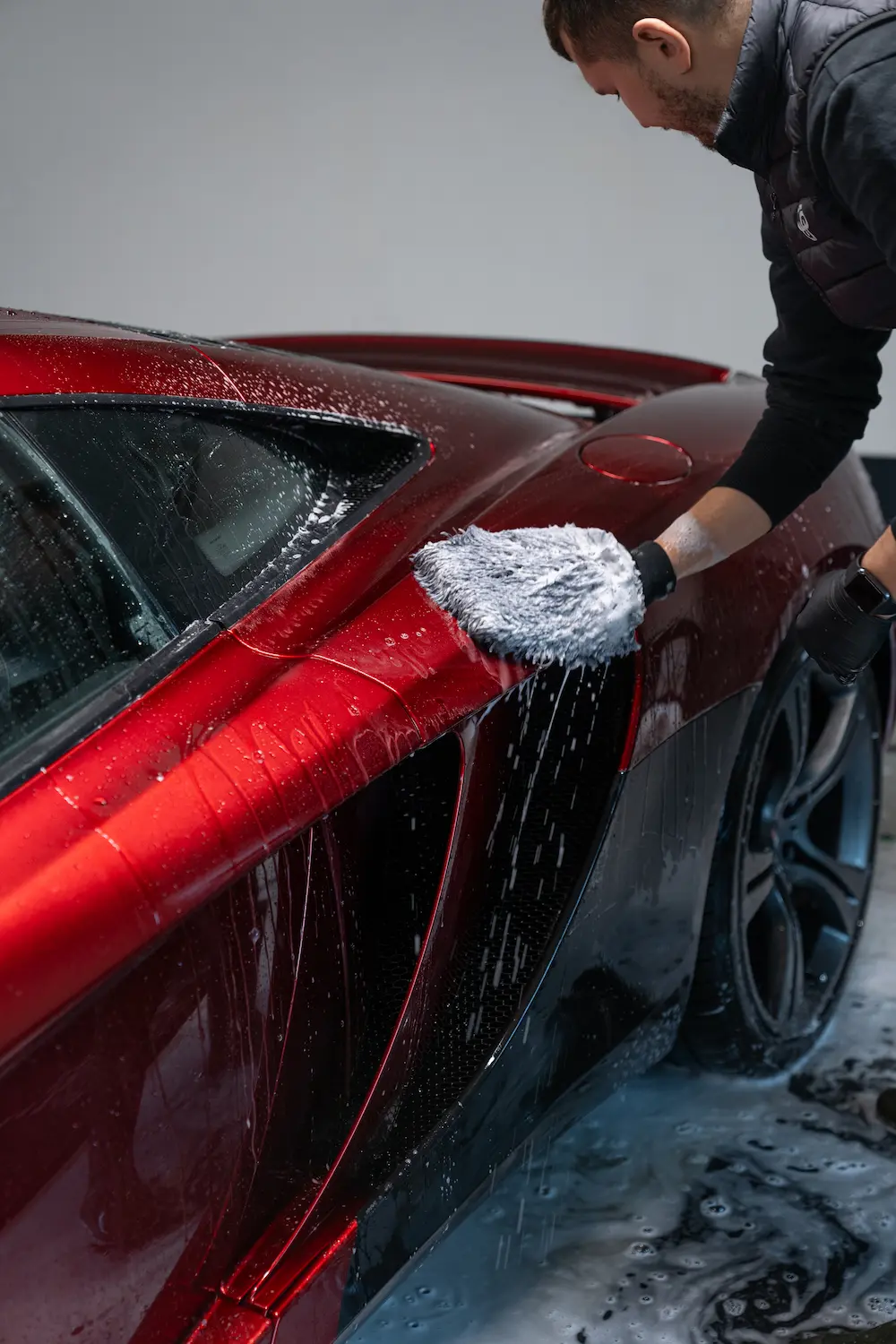 Close-up of a technician hand-washing a red supercar with a foam mitt, water and soap dripping over the bodywork inside a professional detailing workshop.