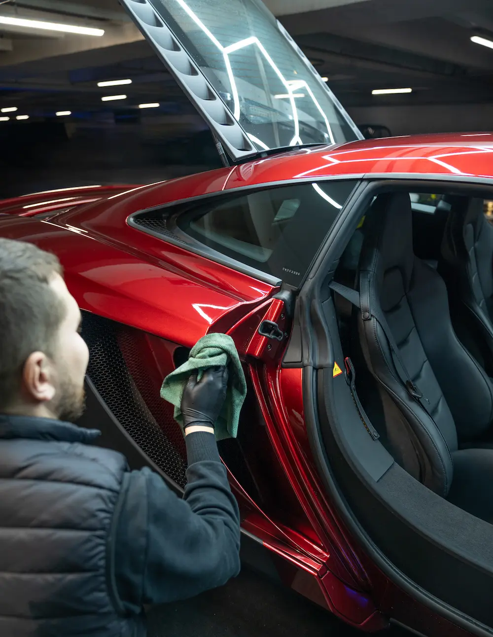 Carsup expert carefully wiping the door frame of a red supercar with a microfiber cloth, door raised, inside a modern automotive detailing workshop.