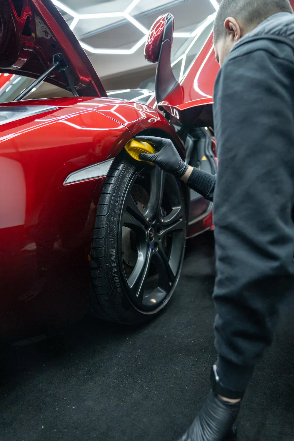 Carsup expert polishing the rear wheel arch of a red supercar with a detailing pad, door raised, inside a modern high-end automotive workshop with geometric ceiling lighting.
