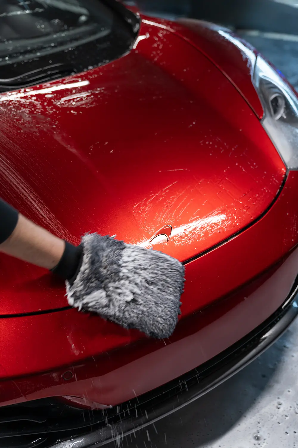 Close-up of a Carsup expert hand-drying the hood of a red supercar with a microfiber towel, water droplets visible on the glossy paint inside a professional detailing workshop.