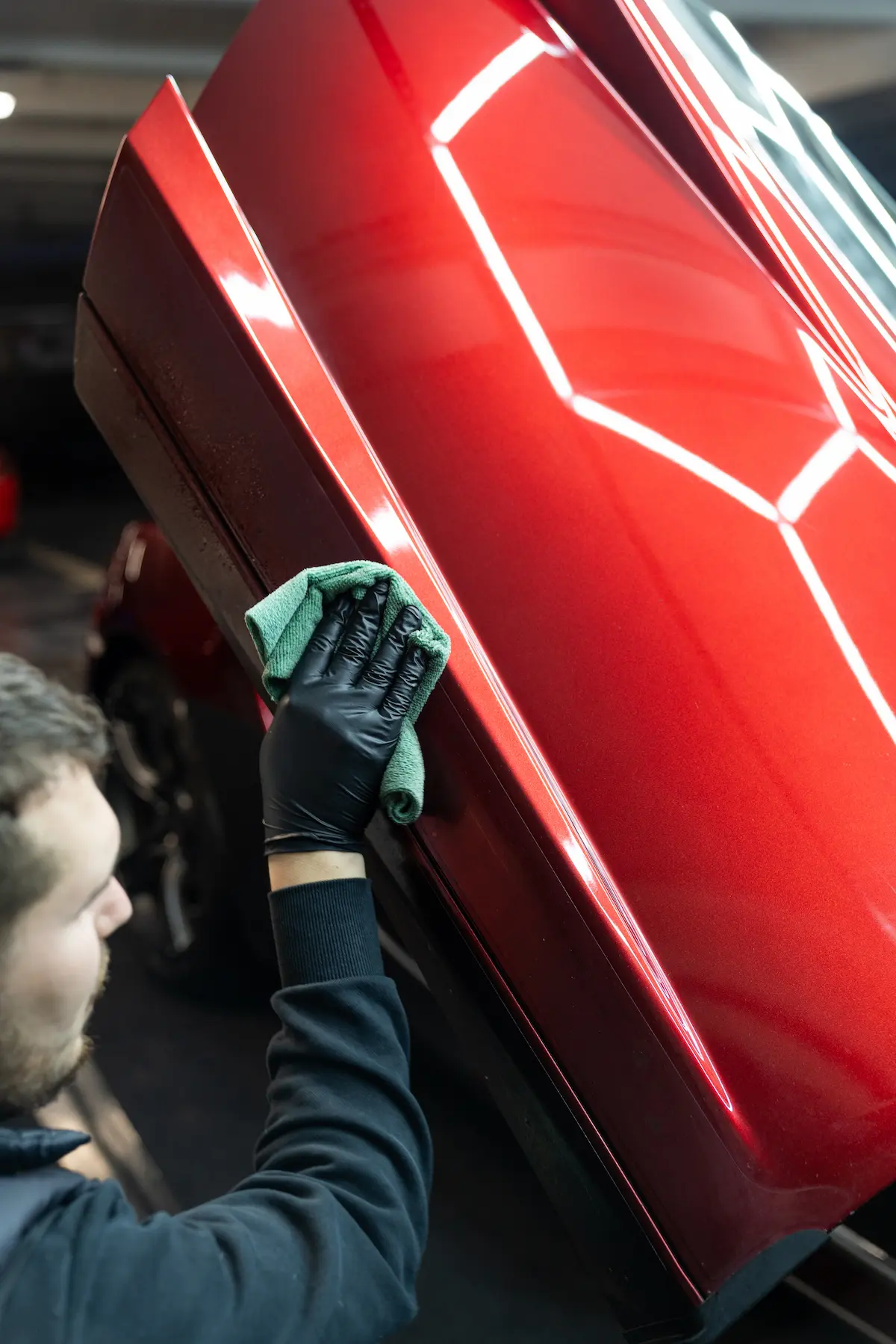 Carsup expert carefully wiping the raised door panel of a red supercar with a microfiber cloth, bright geometric ceiling lights reflecting on the glossy paint inside a high-end detailing workshop.