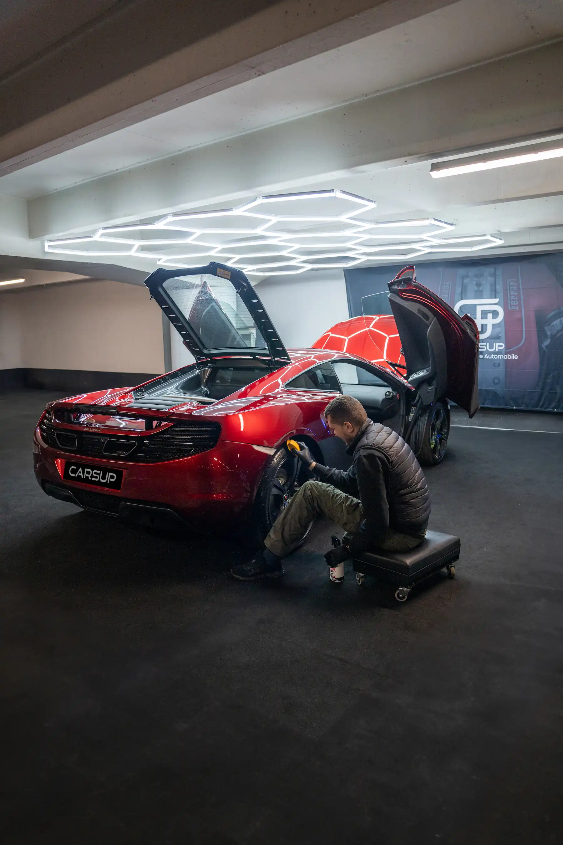 Technician polishing the rear wheel arch of a red McLaren sports car with both doors raised inside a luxury automotive workshop, with a “CARSUP” plate visible.