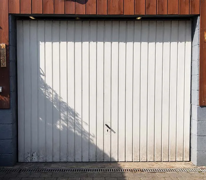 Porte de garage blanche en métal avec ombre et poignée centrale, encadrée de bois et béton.