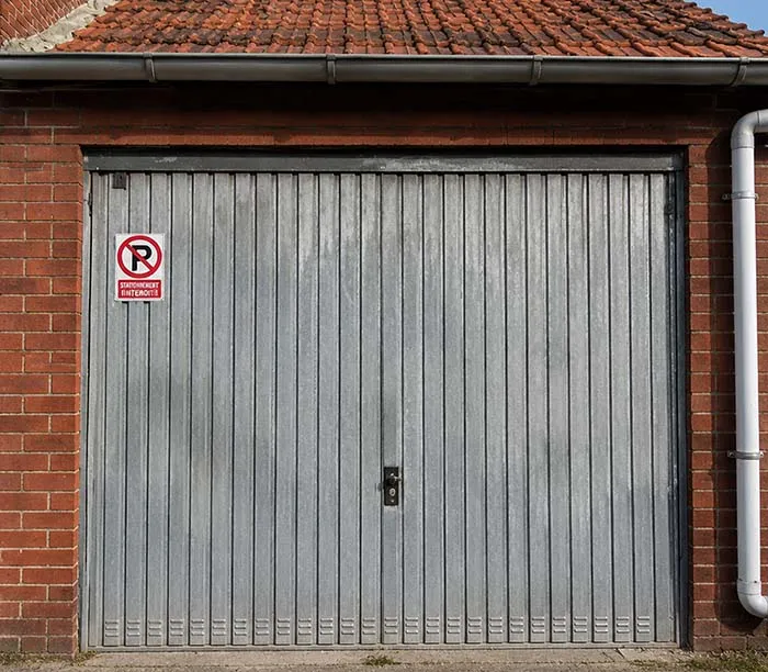 Porte de garage grise en métal avec un panneau indiquant "Stationnement interdit" sur un mur en briques rouges.