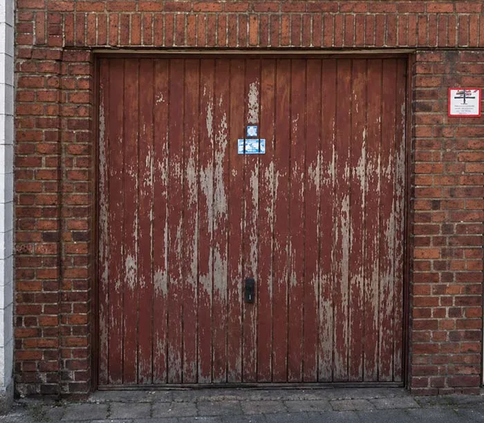 Porte de garage en bois rouge écaillé encadrée de briques rouges avec panneau blanc à droite.