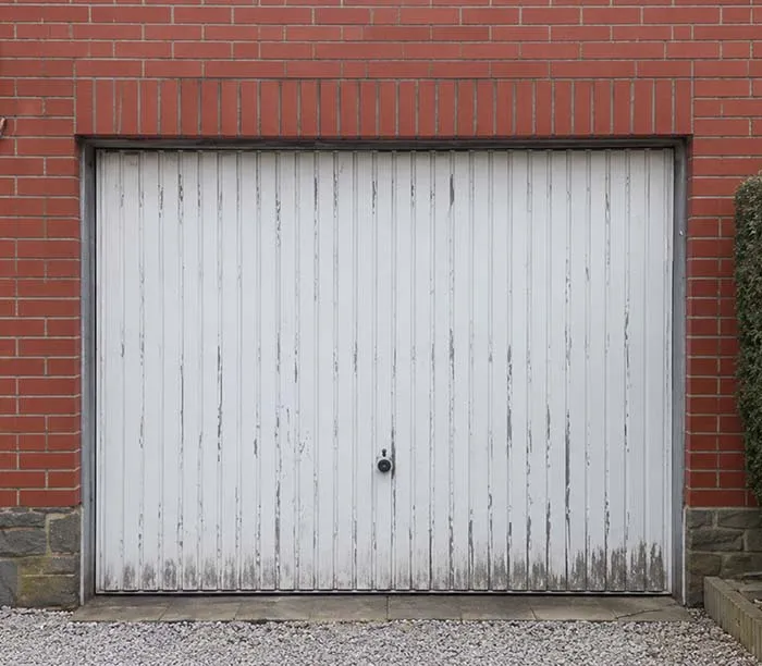 Porte de garage blanche vieillie avec vernis écaillé dans un mur en briques rouges et pierre.