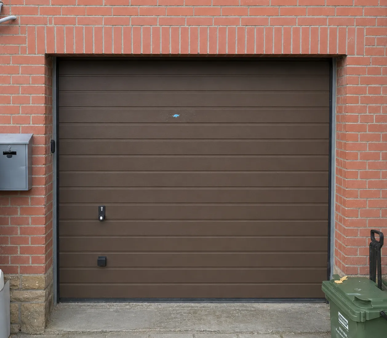 Porte de garage marron avec boîtier de serrure, encadrée de briques rouges et conteneur vert à droite.