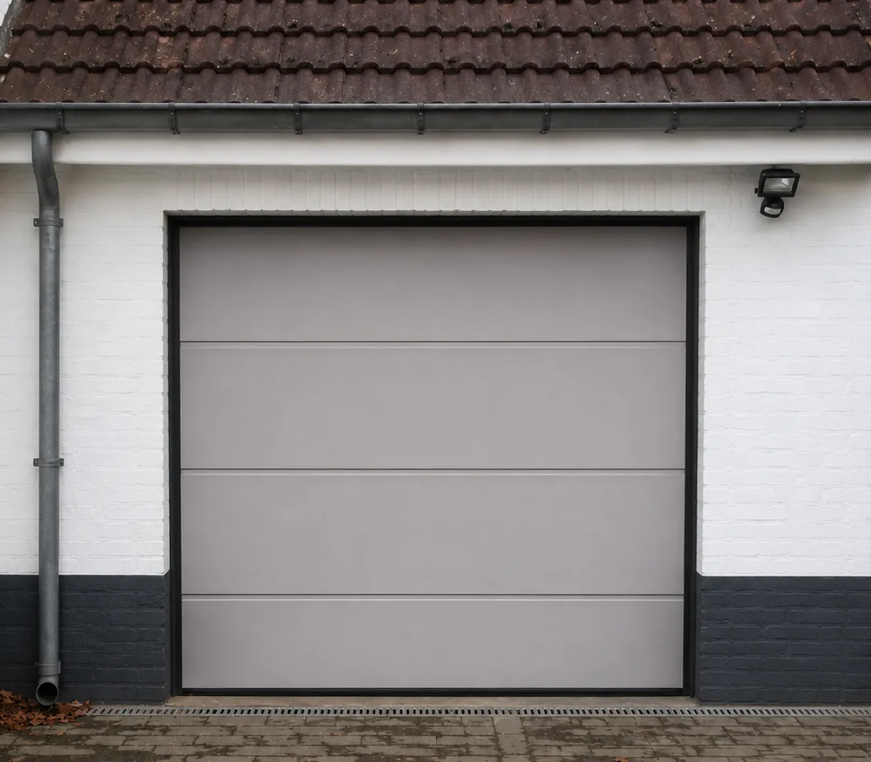 Porte de garage grise moderne encadrée de murs blancs et noirs sous un toit en tuiles marron.