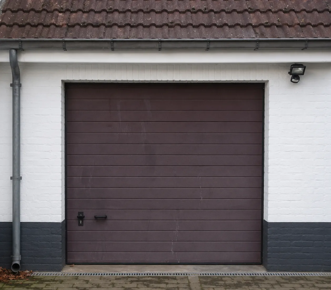 Porte de garage marron fermée sur un mur blanc avec base grise, auvent et gouttière grise.