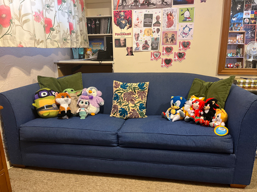 Blue couch with two green pillows and a decorative floral pillow, featuring various plush toys including characters like Minion, owl, purple creature, Sonic, Knuckles, and Tails.