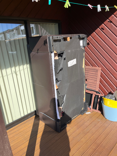 Damaged black refrigerator with broken exterior, wooden chair, and blue bucket.