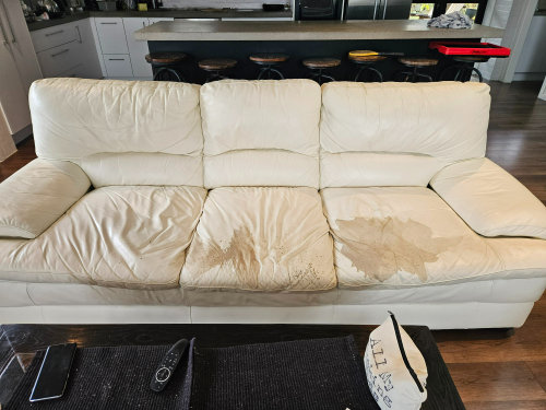 White leather couch with stained cushions, remote control, and decorative pillow.