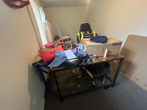 Desk with scattered clothes, a chair, a box, and some papers.