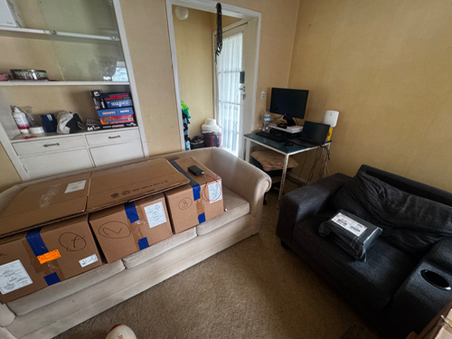 Boxes stacked on a couch, a computer desk with a monitor, and a black sofa.