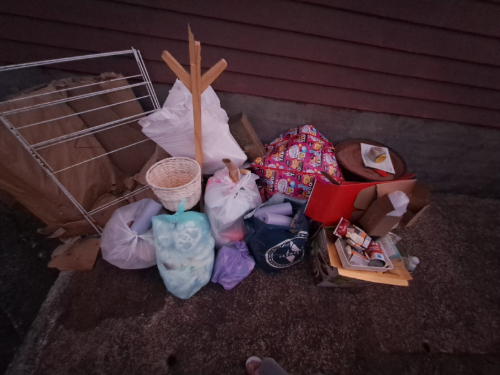 Collection of garbage bags, cardboard boxes, a wicker basket, a large patterned bag, a wooden rack, and various discarded items.