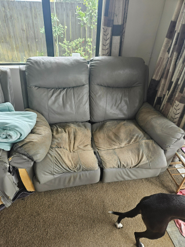 Gray leather reclining loveseat with worn cushions, a blue blanket draped over one arm, and the hind legs of a dog in the foreground.