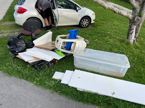 White plastic chair, blue plastic chair, transparent storage box, black garbage bag, wooden boards, white panel, office chair, person unloading items from a car.