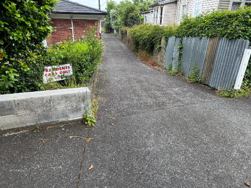 Concrete pathway, brick wall, overgrown bushes, "RESIDENTS CARS ONLY" sign, corrugated metal fence.