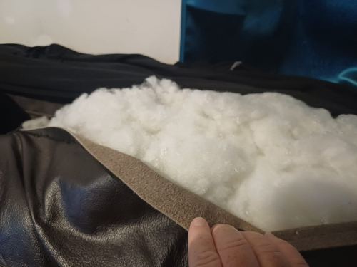 Hand holding back a tan fabric layer revealing white stuffing on a black leather surface.