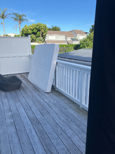 White mattress leaning against a railing, black bean bag chair on wooden deck.