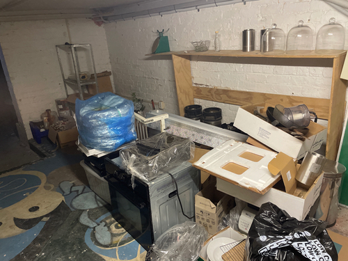 Cluttered room with boxes, a blue tarpaulin, a washing machine, kitchen appliances, glass containers, and a wooden shelf.