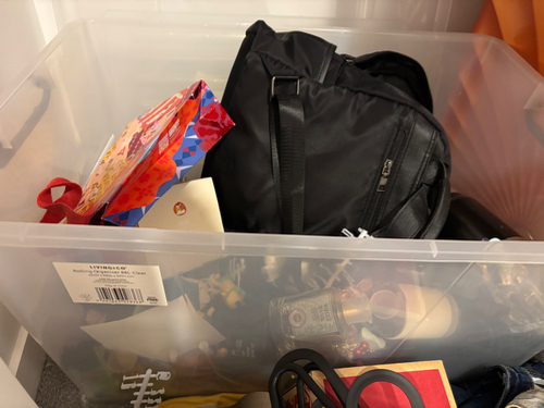 Clear storage bin with a black backpack, a colorful gift bag, and various items including papers and small objects.