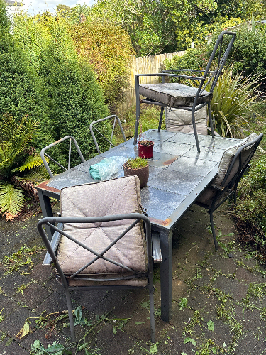 Metal outdoor table with four chairs, one chair stacked on the table, cushion-covered seats, and a small potted plant.