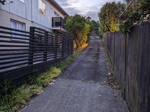 Pathway between black wooden fences lined with grass and plants, leading to a concrete surface.