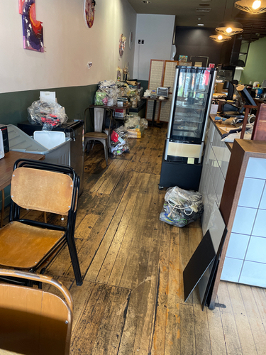 Interior of a cafe with wooden flooring, stacked boxes, an empty display refrigerator, and a few chairs.