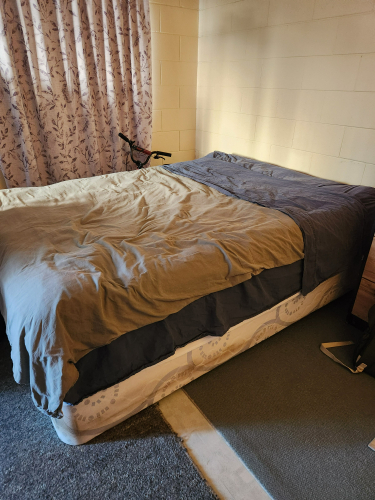 Bed with a gray and beige comforter, bedside table, and bicycle.