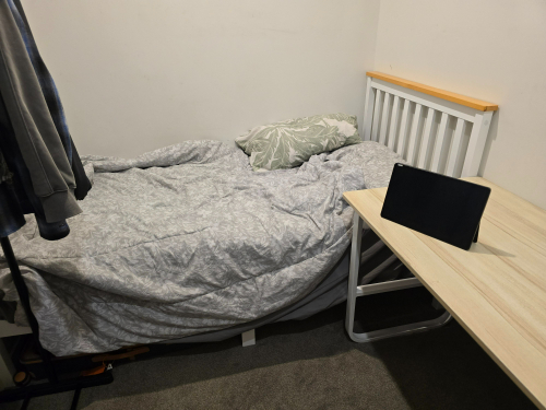 Bedding with a patterned duvet, a green pillow, a wooden bed frame, and a tabletop with a tablet.