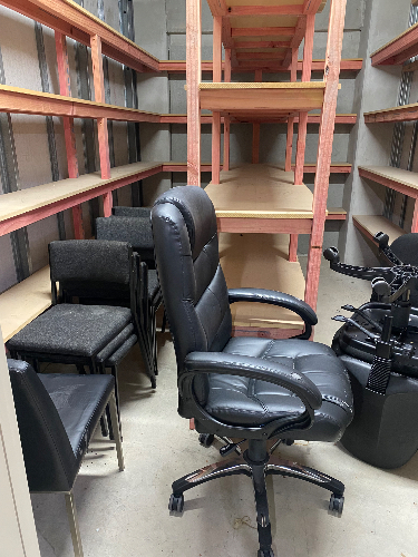 Black ergonomic office chair, stack of metal-framed chairs, wooden shelves.