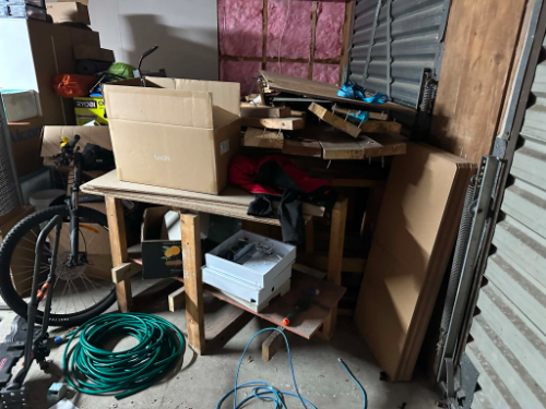 Cluttered table with cardboard boxes, wooden pallets, bicycle, green extension cord, electronic device, and piles of materials.