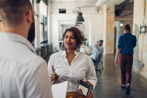 Woman in white shirt holding a notebook discussing with bearded man in an office space.