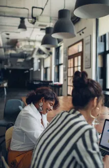 Two women working together in a modern office, one smiling and looking down while the other focuses on a laptop.