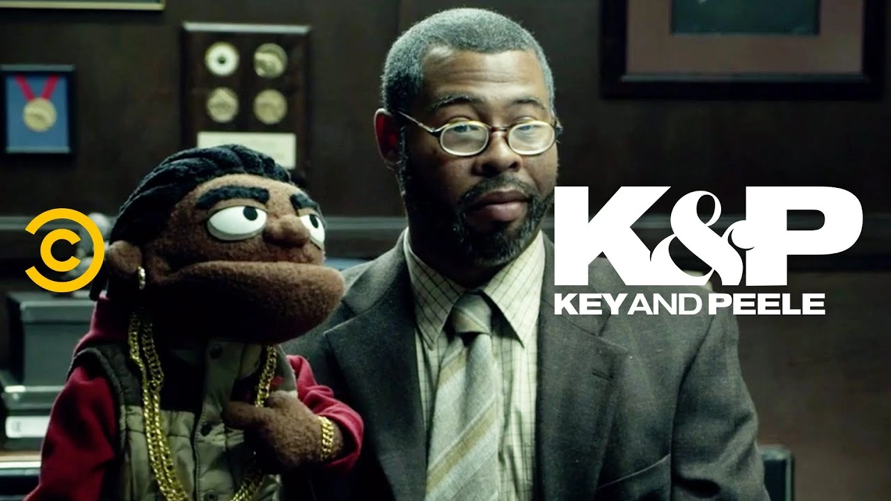 Source: Key and Peele's YouTube thumbnail. A man in glasses sits beside a puppet wearing a red jacket and gold chains, with the text "K&P KEY AND PEELE" displayed on the right.