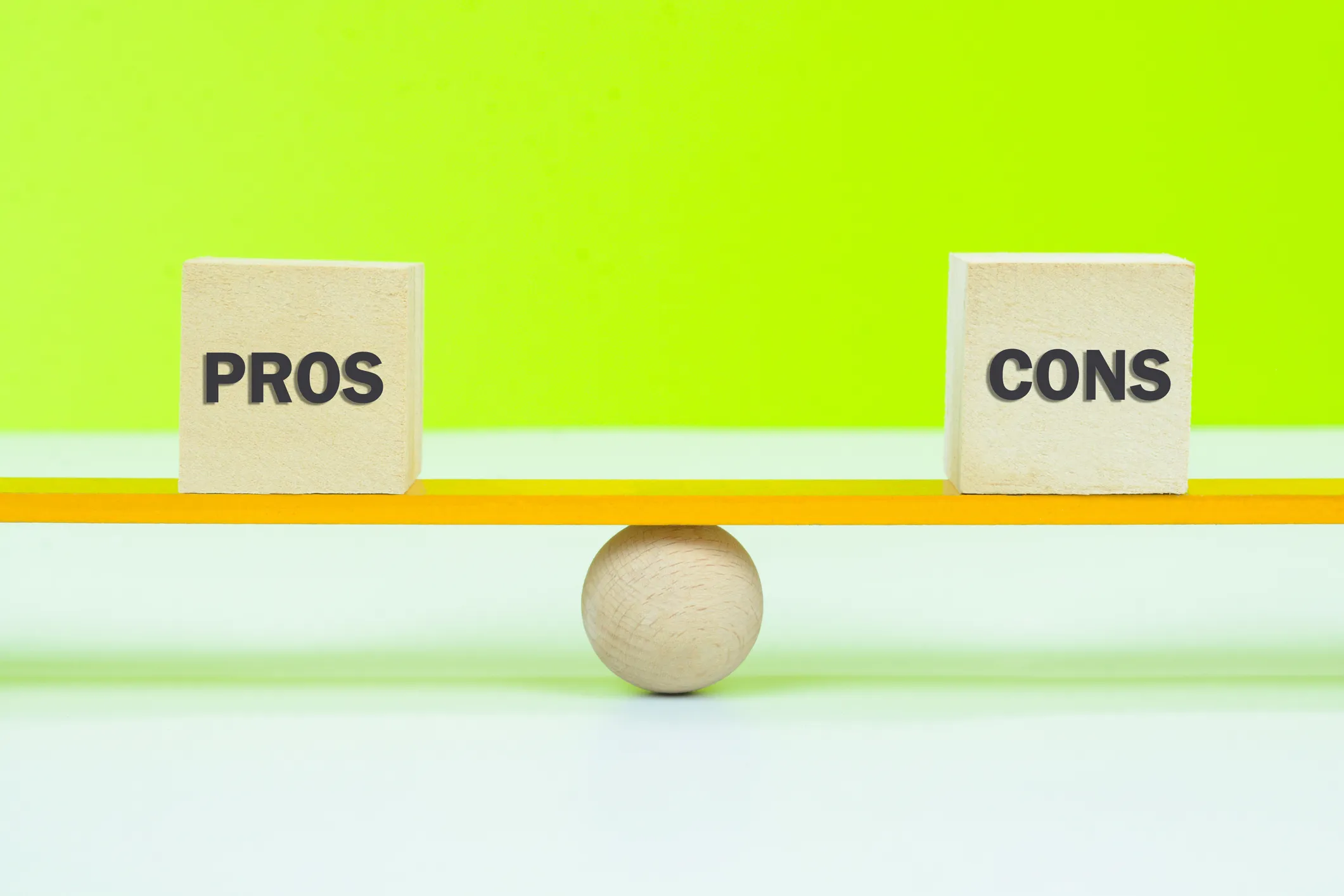 Wooden blocks labeled "Pros" and "Cons" balanced on a seesaw, representing decision-making.