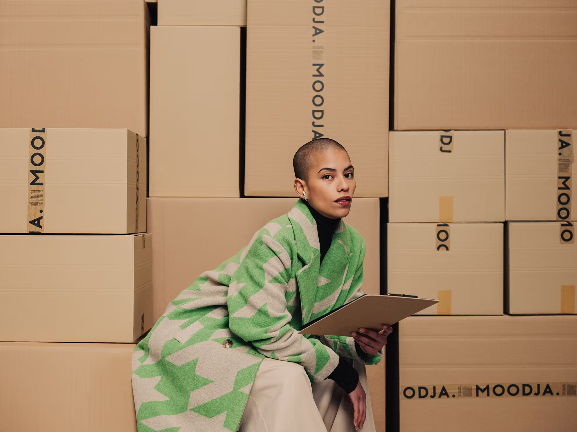 fulfillment mode, model mit grüner jacke sitzt zwischen moodja paketen und hält klemmbrett