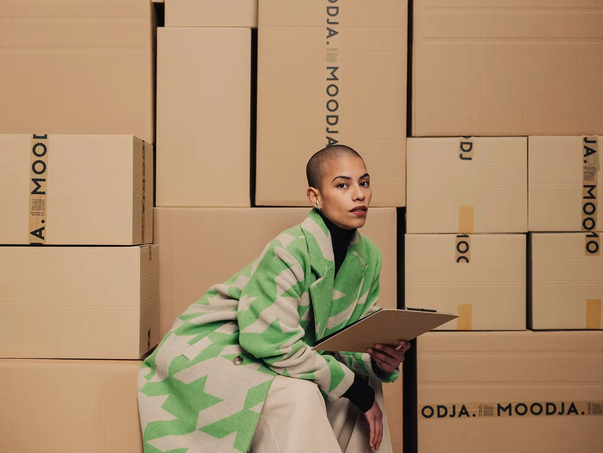 fulfillment mode, model mit grüner jacke sitzt zwischen moodja paketen und hält klemmbrett