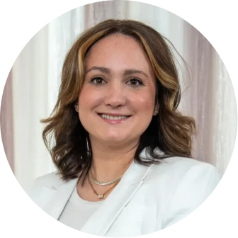 Smiling woman with shoulder-length wavy hair wearing a white blazer and layered necklaces against a light neutral background.